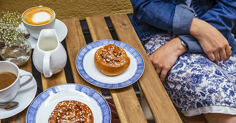 Fika w Sztokholmie: Dwie filiżanki kawy, dzbanuszek mleka i bułeczki cynamonowe (kanelbullar) na drewnianym stoliku na zewnątrz.