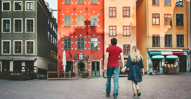 Para trzymająca się za ręce spaceruje po brukowanym placu Stortorget w Sztokholmie, otoczonym kolorowymi zabytkowymi kamienicami.