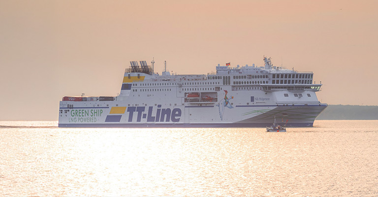 Prom TT-Line 'Green Ship' z napisem 'LNG Powered' płynący po spokojnym morzu o zachodzie słońca, z małą łodzią rybacką w pobliżu.