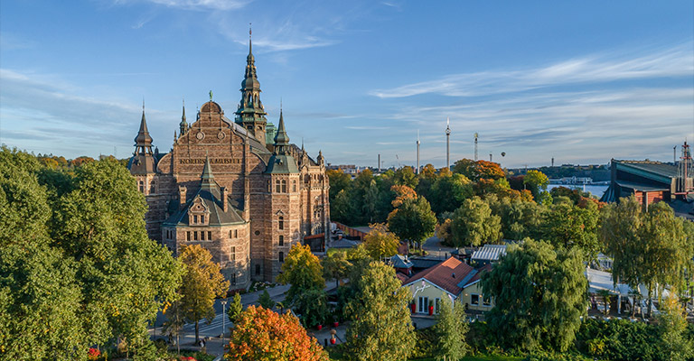 Panoramiczny widok na Muzeum Nordyckie (Nordiska Museet) w Sztokholmie, otoczone jesiennymi drzewami i innymi budynkami pod błękitnym niebem.