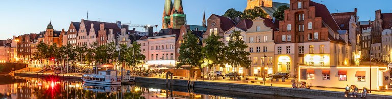 Kvällsutsikt över Lübecks gamla stad med upplysta byggnader och kyrktorn speglade i vattnet.