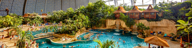 Inomhusmiljö med tropiskt tema – familjer och besökare badar, solar och promenerar bland palmer, solstolar, semesterstugor och pooler i äventyrsbadet Tropical Islands, under en stor glaskupol.